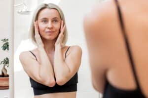 Middle-aged woman touching her face in the mirror and considering a mini or full facelift