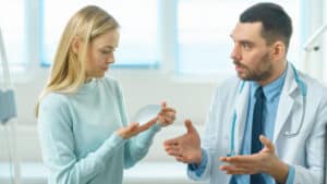 Cosmetic surgeon showing female patient breast implant samples while discussing breast implant replacement surgery in modern clinic